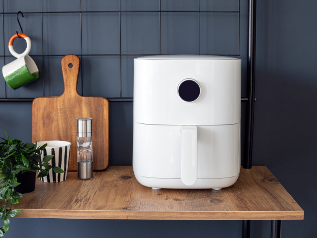 A white air fryer displayed on a wooden kitchen shelf, next to a plant and kitchen accessories.