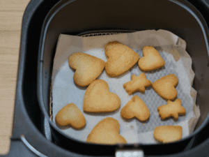 Heart-shaped and star-shaped cookies baking inside an air fryer basket, lined with parchment paper.