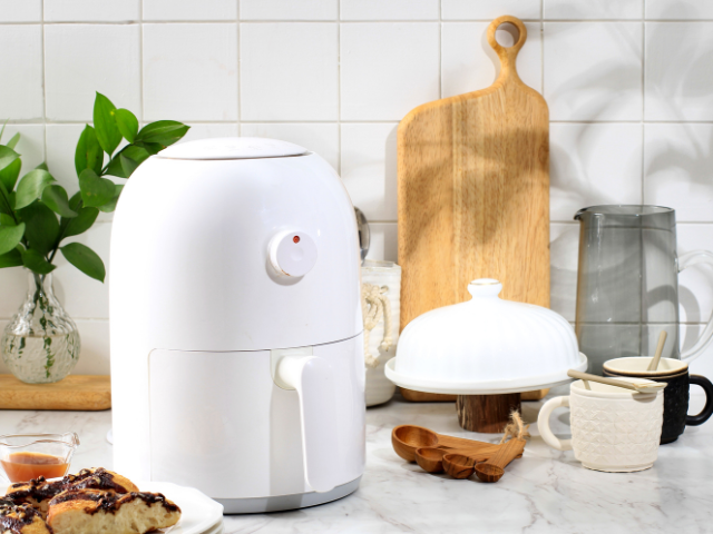 A white air fryer displayed in a cozy kitchen setting with wooden utensils and a plate of pastries.