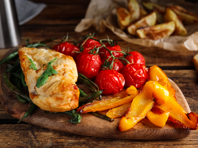 Grilled chicken breast, roasted tomatoes, peppers, and green beans on a wooden serving board.