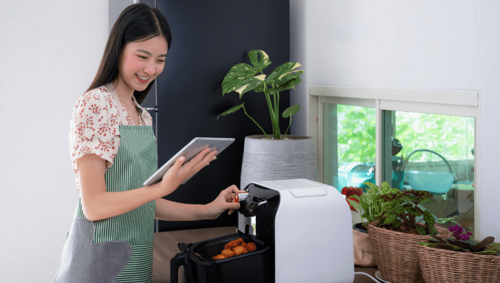 A woman in a kitchen using an air fryer while checking a recipe on a tablet.