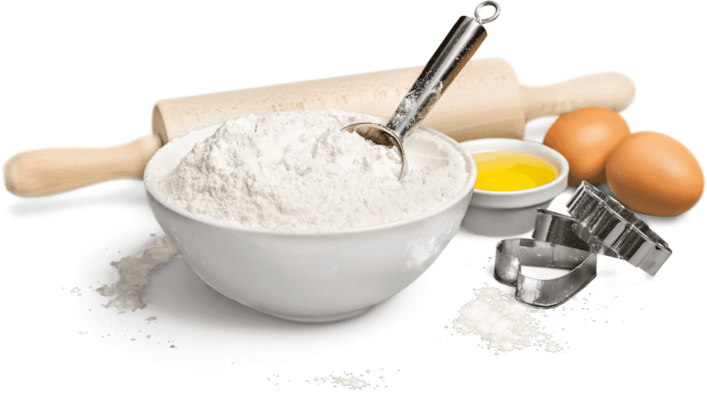A bowl of flour with a measuring spoon, a rolling pin, and eggs on a white surface.