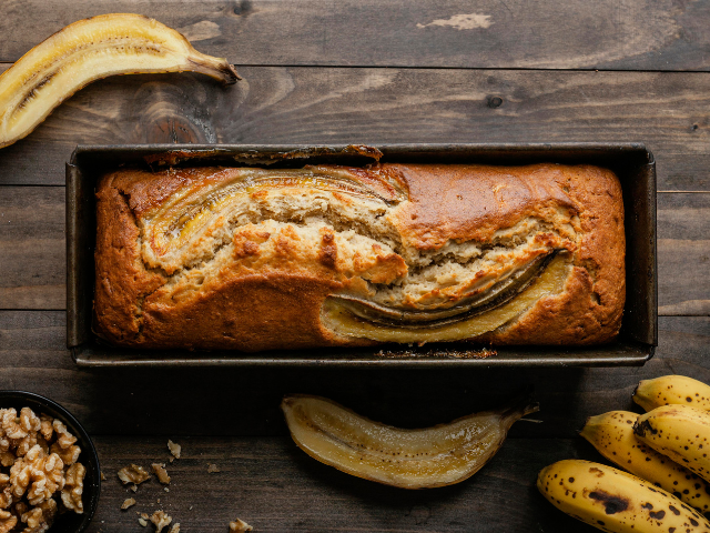 A loaf of banana bread in a tin with ripe bananas and walnuts arranged beside it on a wooden surface.