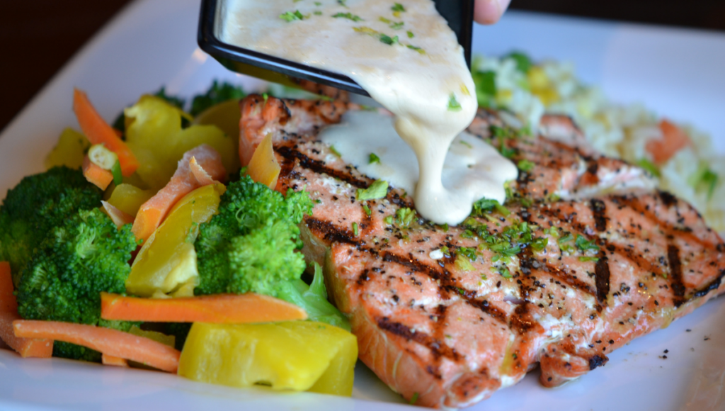 A grilled salmon fillet topped with a creamy sauce being poured over it, accompanied by a colorful array of steamed vegetables, including broccoli, carrots, and yellow squash.