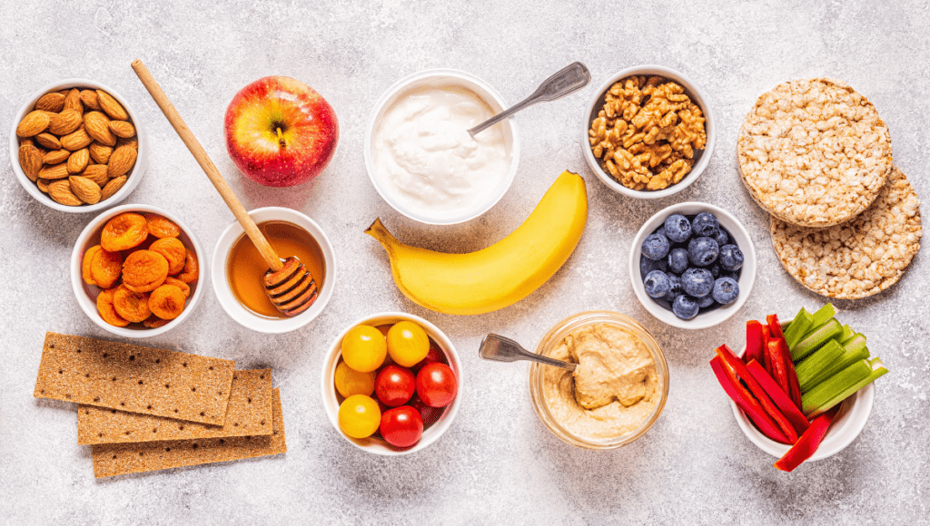 A top-down view of a variety of healthy snacks, including nuts, fruits, hummus, and rice cakes, arranged in bowls on a textured background.