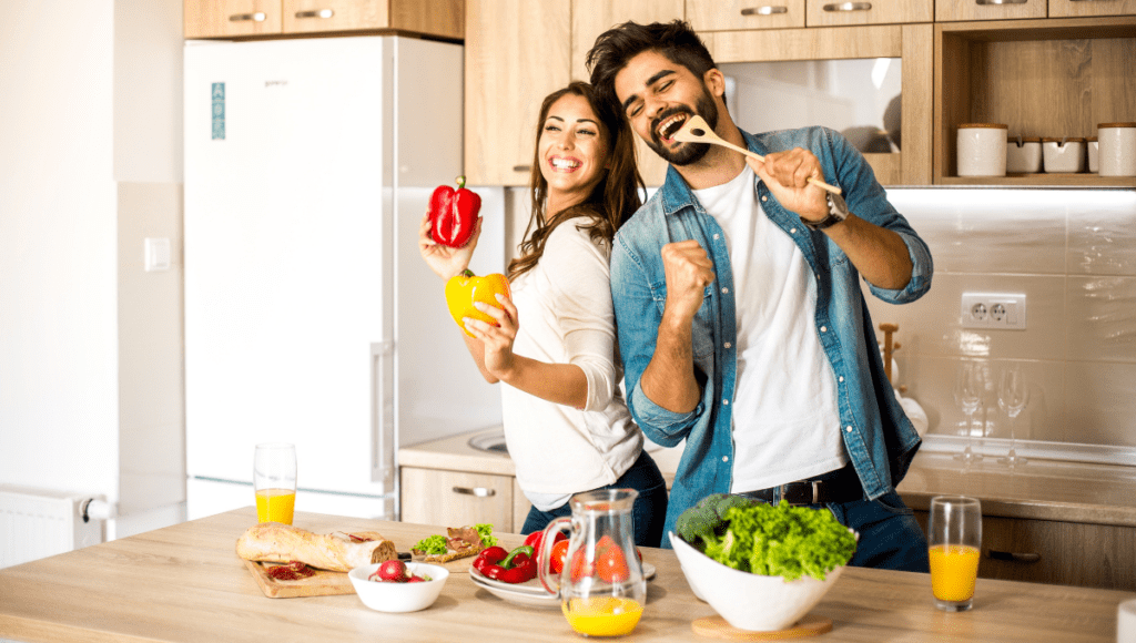 A couple having fun in the kitchen, holding red and yellow bell peppers while dancing.