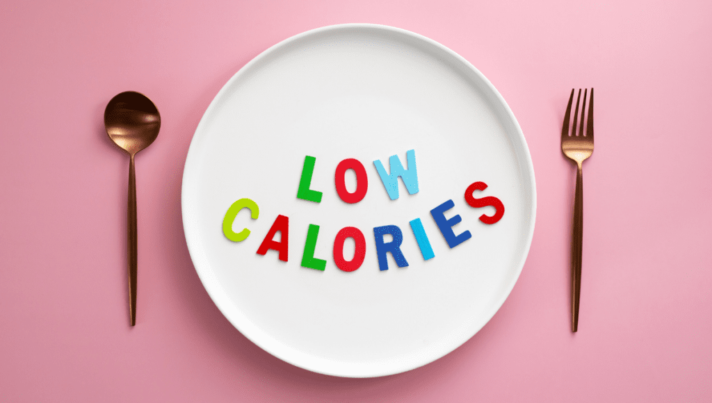 A white plate with colorful letters spelling "LOW CALORIES," placed on a pink background with gold-colored utensils beside it.