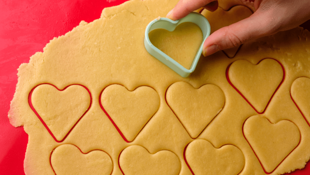 A hand is cutting heart-shaped cookies from rolled-out dough using a cookie cutter. The cookies are arranged neatly on a red background.