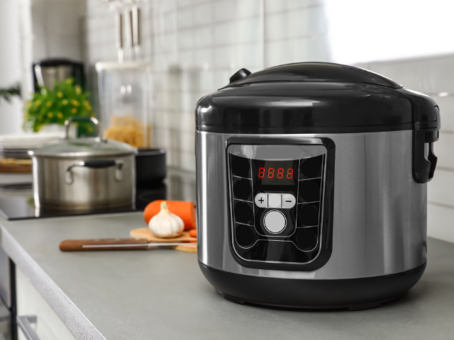 A digital rice cooker sitting on a kitchen counter with other cookware in the background.