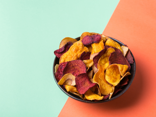 A bowl of vibrant vegetable chips, including beet and sweet potato, against a split green and orange background.