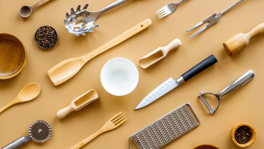 Overhead view of various kitchen utensils, including wooden spoons, a knife, and a bowl, arranged on a beige background.
