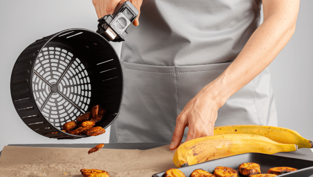 A person using an air fryer to pour out crispy plantains onto parchment paper.