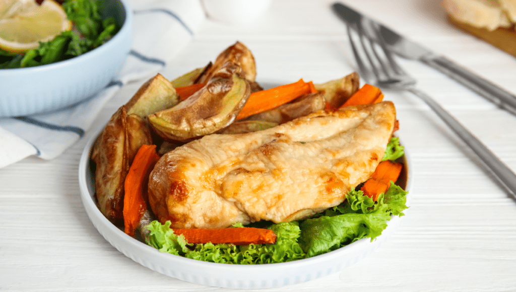 A plate of baked chicken breast served with roasted potato wedges, carrots, and salad.