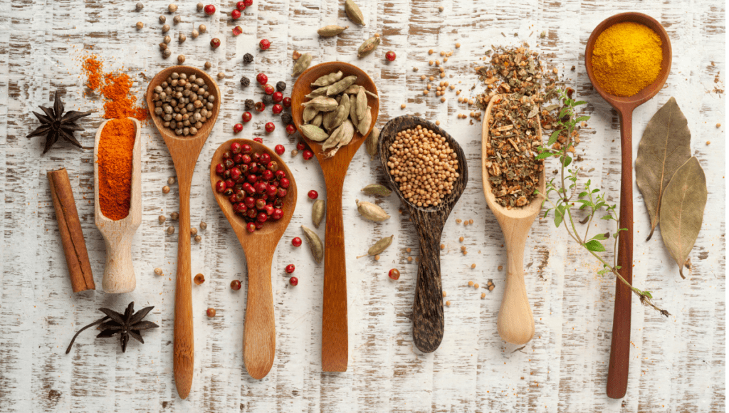A variety of spices beautifully displayed in wooden spoons, featuring ingredients like peppercorns, mustard seeds, bay leaves, and chili powder.
