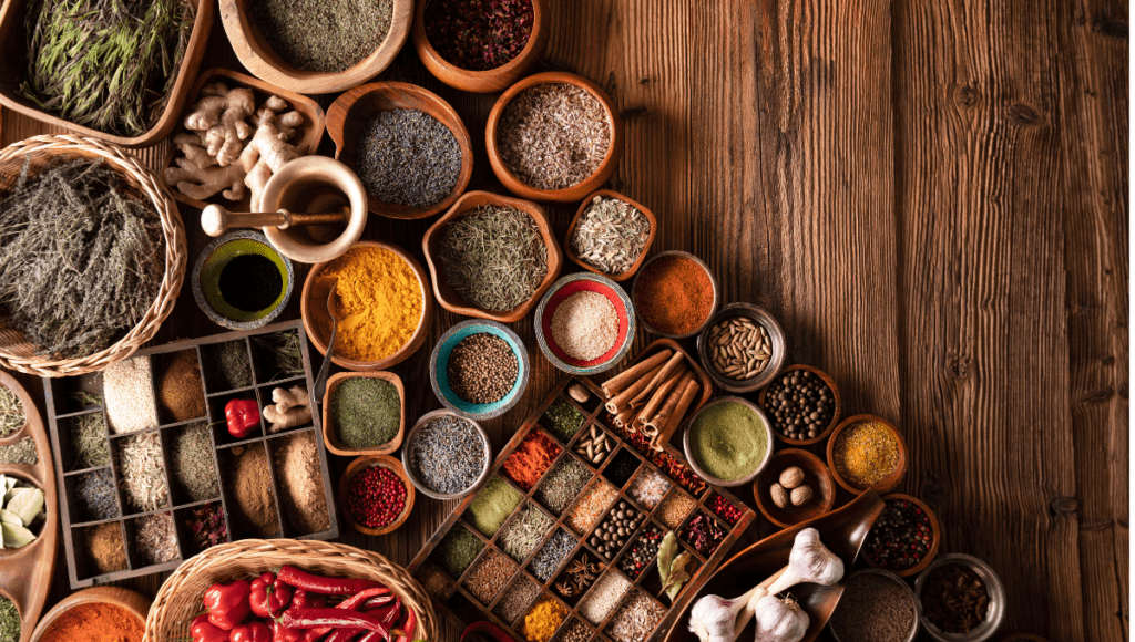 An overhead view of various dried herbs and spices in wooden bowls, arranged neatly on a rustic wooden table.