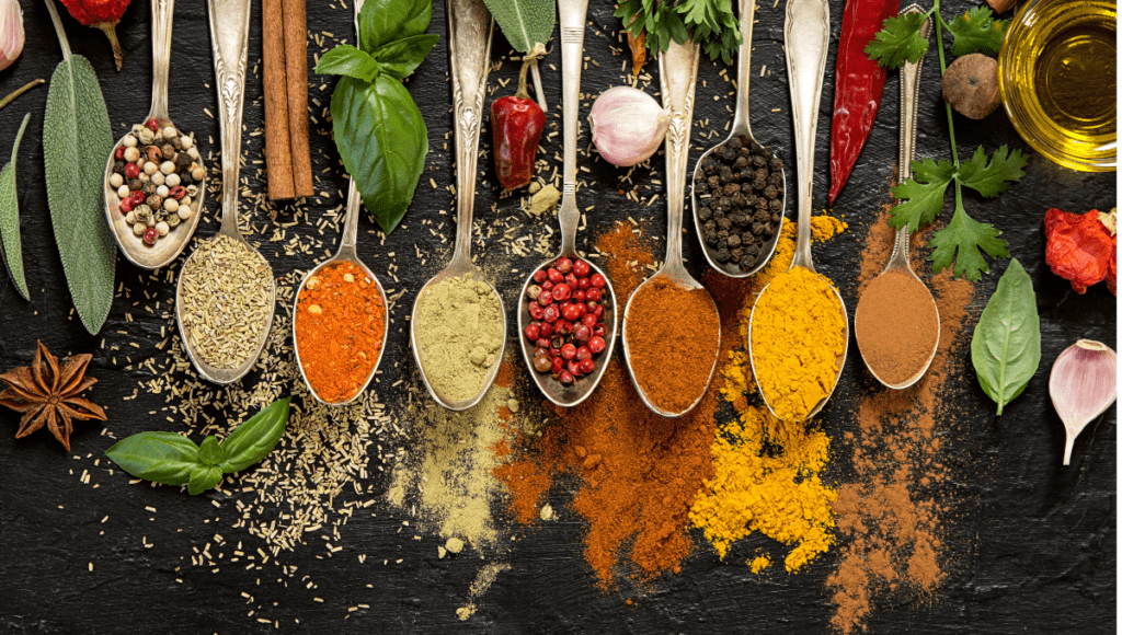 A variety of herbs and spices on spoons, including peppercorns, cinnamon, and basil leaves, against a black background.