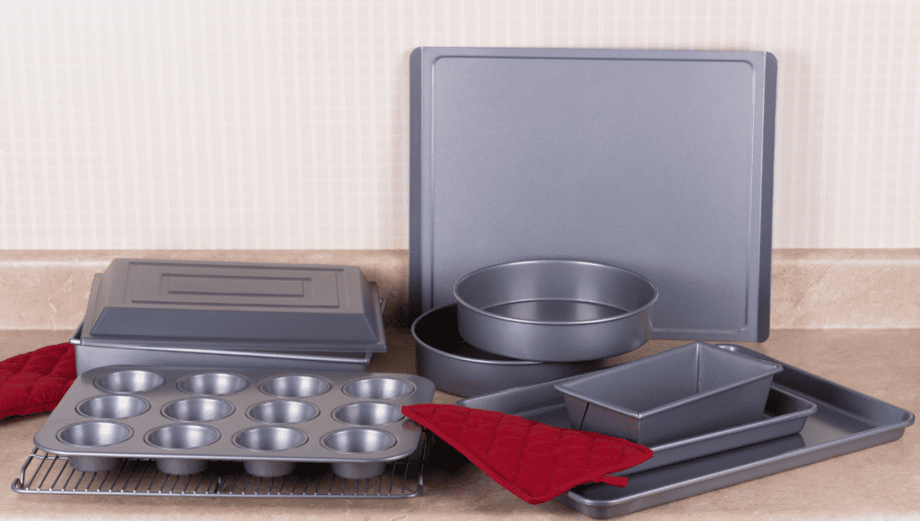 A set of non-stick bakeware, including muffin pans, round pans, and baking trays, arranged neatly on a kitchen countertop with red oven mitts.