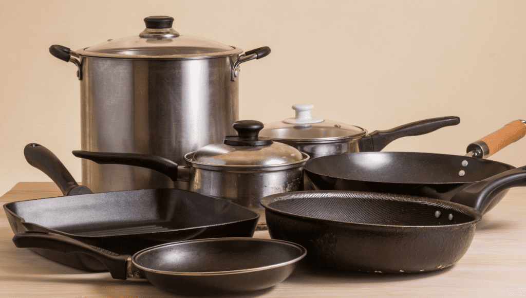 A collection of various pots and pans, including a large stainless steel stockpot, frying pans, and a saucepan with lids, set on a light wooden surface.