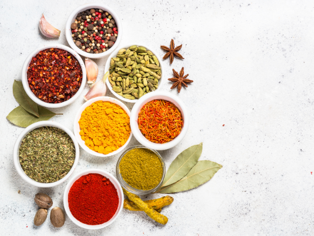A colorful assortment of spices in various bowls, including peppercorns, cardamom, and turmeric, displayed on a white textured surface.