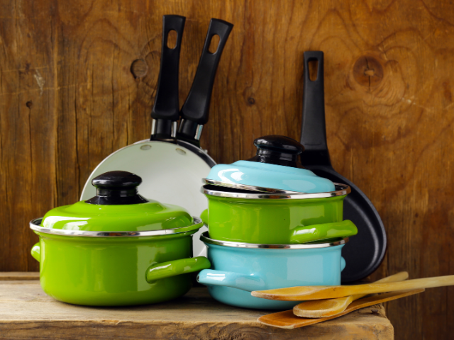 A colorful set of pots in green and blue with black handles, accompanied by wooden cooking utensils, arranged against a rustic wooden backdrop.