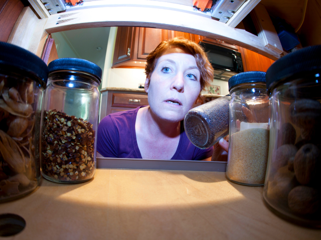 A woman with a surprised expression, peeking into a pantry shelf filled with jars of spices and dried goods.