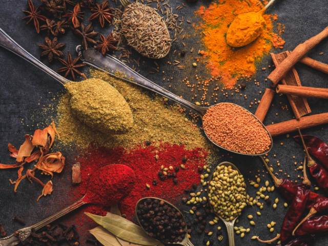 Several spoons filled with different spices, including turmeric, red chili powder, cumin, and coriander, laid out artistically on a dark background.