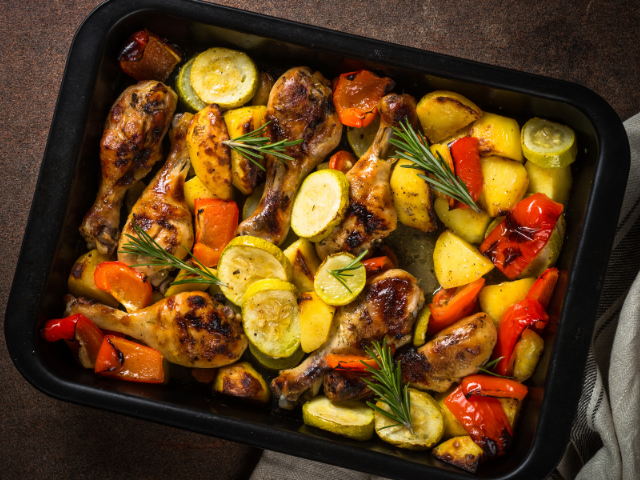A tray of roasted chicken drumsticks with zucchini, bell peppers, and potatoes, seasoned with fresh herbs.